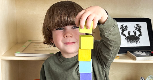 Boy building a tall tower with vibrant wooden blocks, with Quiggly’s magnetic maze and high-contrast baby toy set visible on the shelf in the background