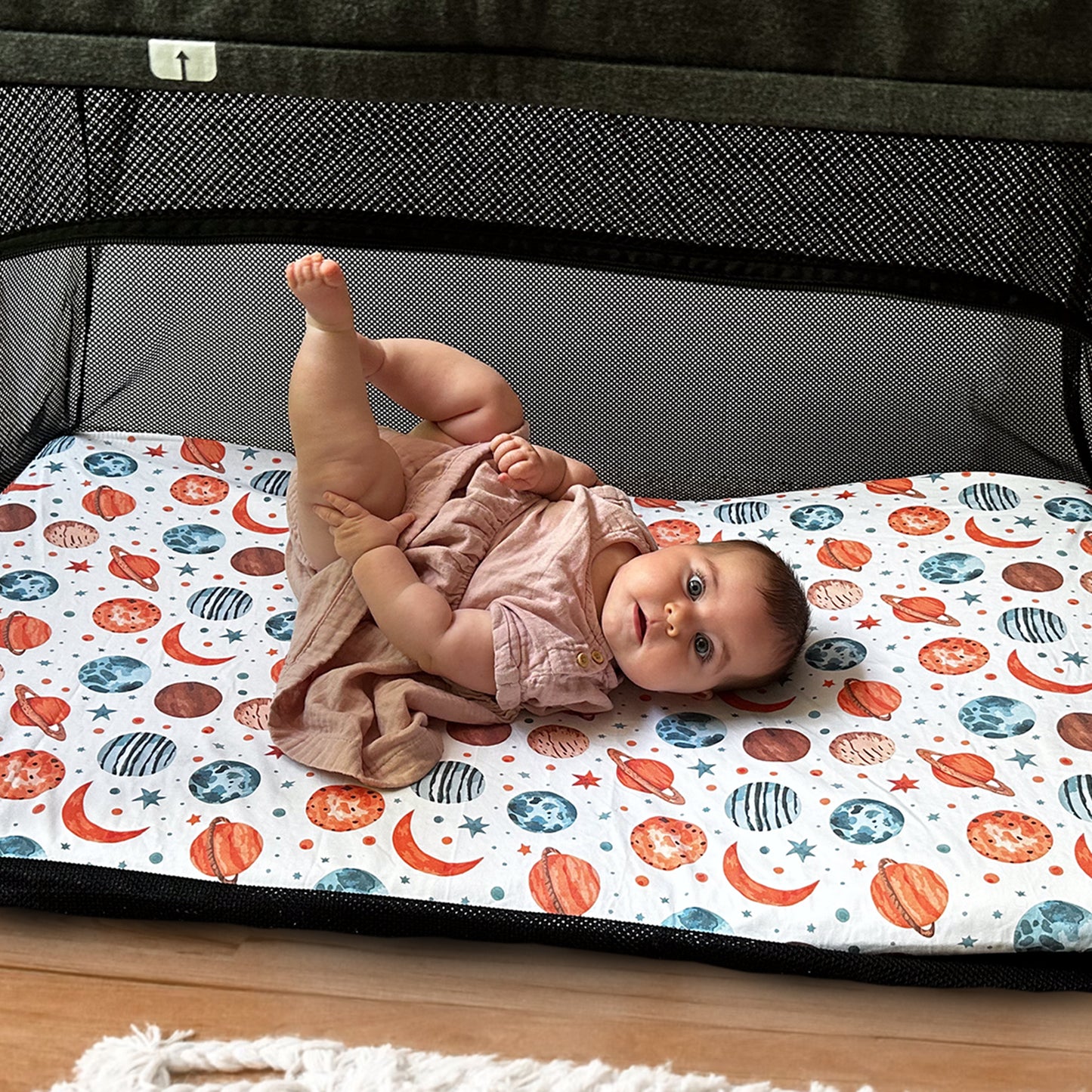 Curious baby lying on planet-patterned travel crib sheet inside Guava Lotus travel crib