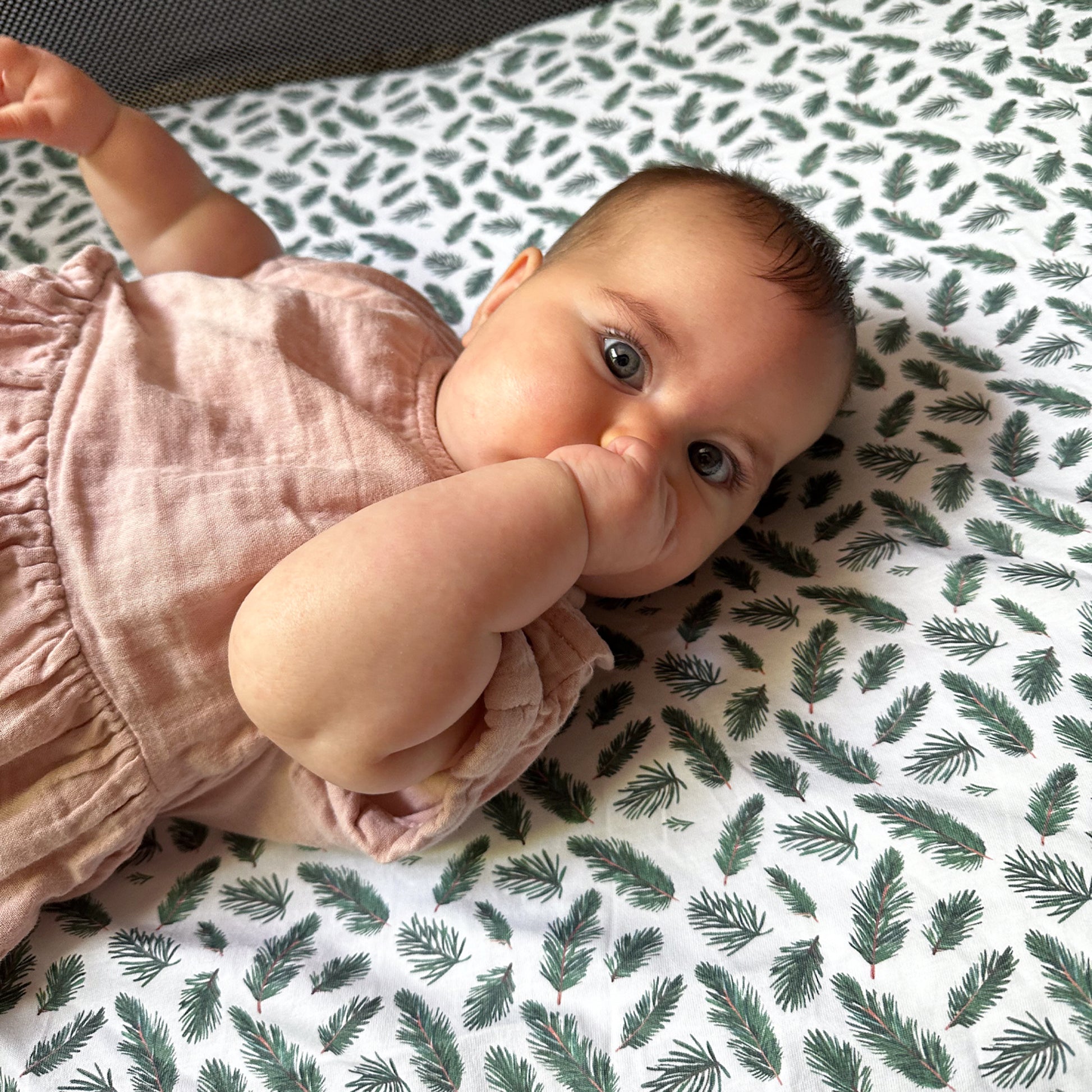 Smiling baby lying on leaf-patterned travel crib sheet inside Guava Lotus travel crib