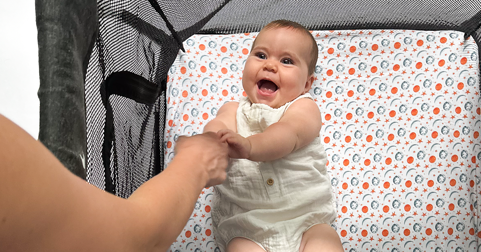 Happy baby clutching her mother’s hand in a guava lotus travel crib covered by Quiggly’s beautifully illustrated planet-themed sheet