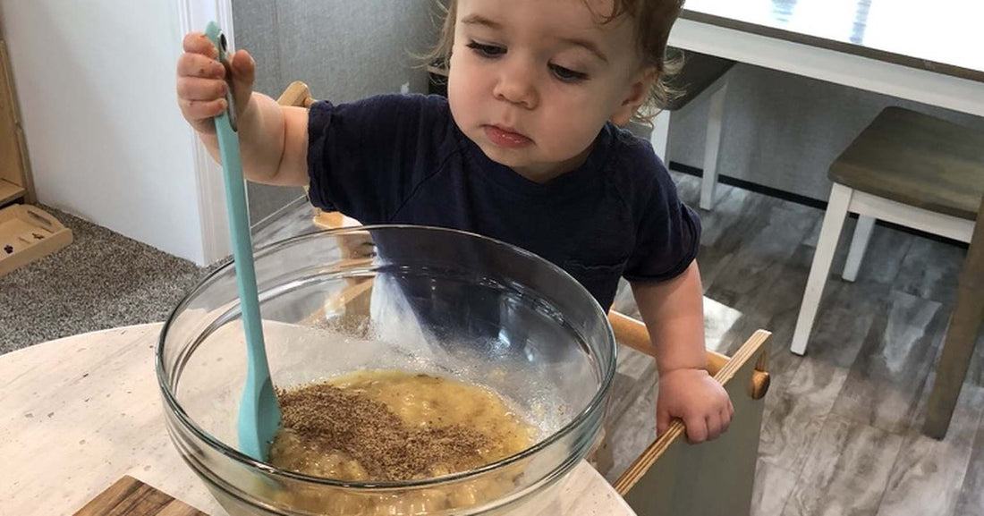 Toddler mixing batter with a spatula while standing in a wooden kitchen helper tower