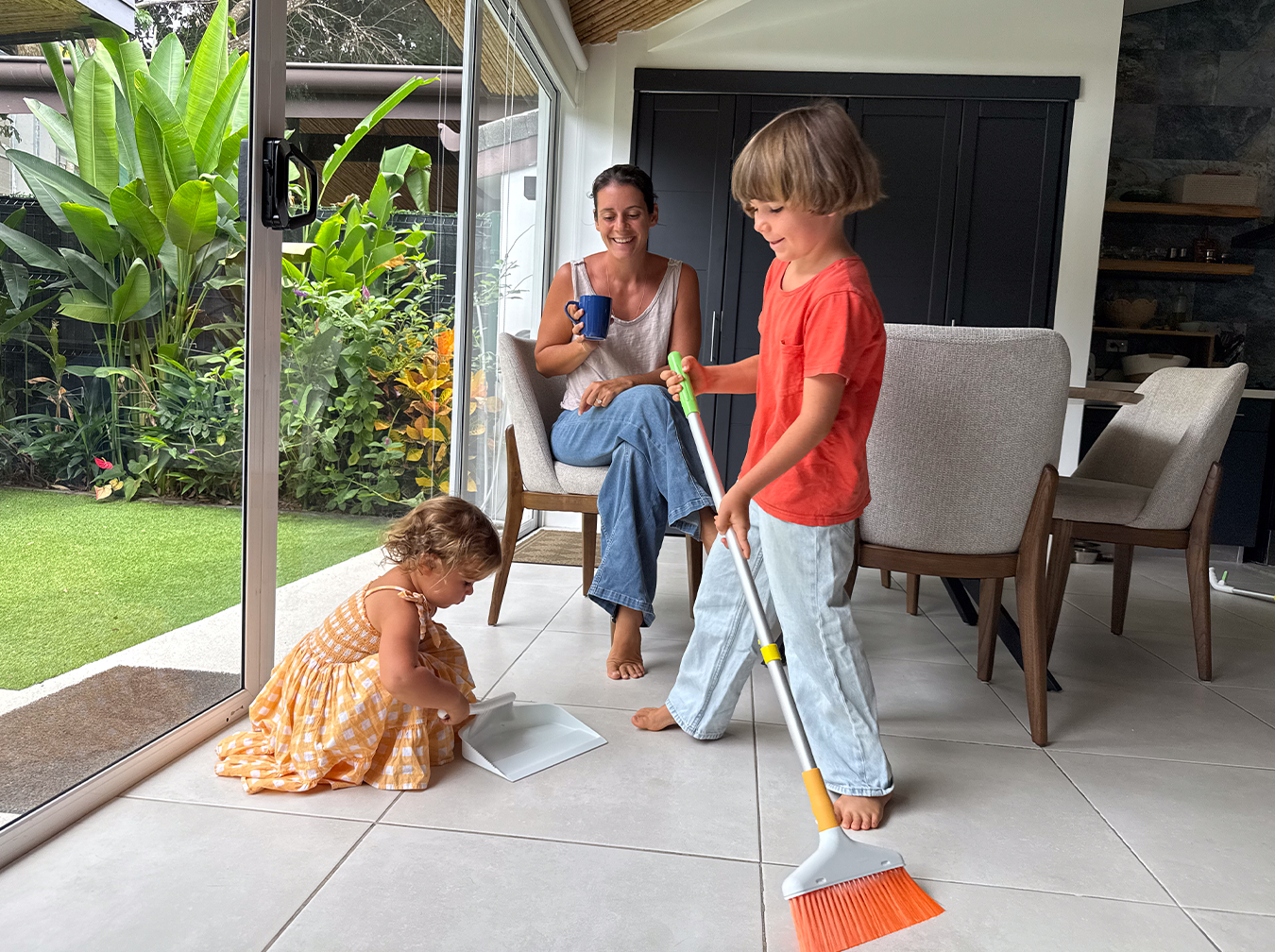 Quiggly founder watching 3 year old use small brush and dustpan while 7 year old sweeps with kids broom from Quiggly cleaning set