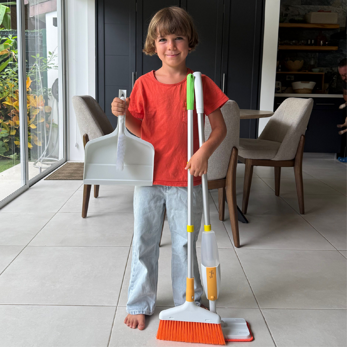 7 year old child holding Quiggly big kids cleaning set with real spray mop, broom, dustpan, and small brush